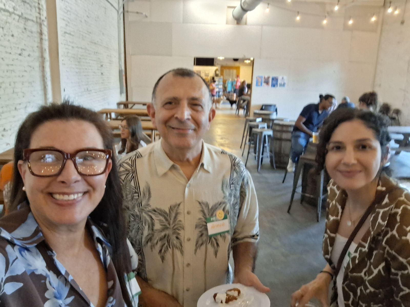 Three attendees at the launch meetup, one holding a plate of food, with the brewery's tables behind them.