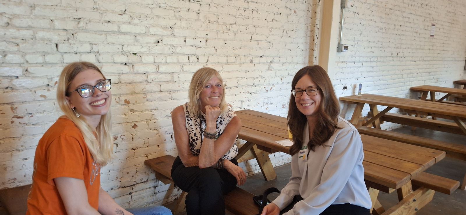 Three attendees seated at picnic tables against the brewery's white brick wall.